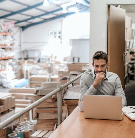 Manager working on a laptop while sitting at a desk in a carpet warehouse with shelves of stock in the background
